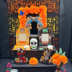 May include: A three-tiered black wooden altar display featuring orange marigold flowers, butterfly accents, two picture frames with skull and cat motifs, a central skull figurine, two skeleton figures, and a glass vase with orange flowers.