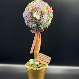 May include: A decorative Easter egg topiary in a gold pot. The egg is adorned with colorful flowers, beads, and two white bunny figures. A pink ribbon and a small sign with text are attached. The pot contains moss and decorative eggs.