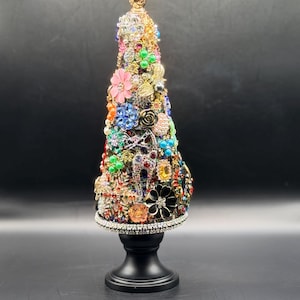 Op de afbeelding: Een decoratieve kerstboom gemaakt van diverse kleurrijke juwelen en ornamenten. De boom is versierd met een verscheidenheid aan vormen en kleuren, waaronder roze bloemen, blauwe edelstenen en gouden accenten. Hij staat op een zwarte sokkel.