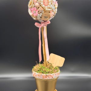 Puede incluir: Un topiario decorativo en una maceta dorada. La parte superior esférica está adornada con flores rosas, perlas y joyas vintage. Una cinta rosa y un palo de madera sostienen el topiario. La maceta está llena de musgo y pequeñas piedras.