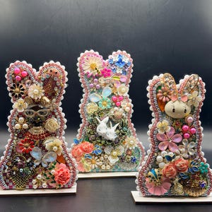 Op de afbeelding: Drie decoratieve konijnenbeeldjes. Elk konijn is versierd met een reeks kleurrijke kralen, parels en bloemversieringen. De konijnen zijn gemonteerd op kleine, rechthoekige houten voetstukken, wat een ambachtelijke esthetiek laat zien.