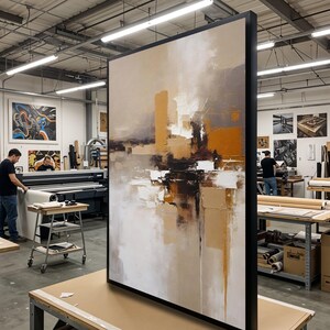 Op de afbeelding: Groot ingelijst abstract schilderij met een zwart frame. Het kunstwerk heeft een palet van beige, bruin, wit en oranje tinten. Het schilderij wordt tentoongesteld in een werkplaats, met ander kunstwerk en apparatuur op de achtergrond.