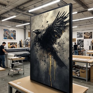 May include: Large framed artwork featuring a black raven with outstretched wings against a textured, gray and gold background. The raven's feathers are detailed, and gold accents drip down. The artwork is displayed in a workshop setting.