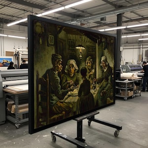 Könnte beinhalten: Großer gerahmter Druck eines Gemäldes, das eine Familie zeigt, die um einen Tisch sitzt, der von einer Hängelampe beleuchtet wird. Das Kunstwerk verwendet gedämpfte Grün-, Braun- und Gelbtöne mit einem schwarzen Rahmen. Das Gemälde steht auf einem Rollständer in einer Werkstatt.
