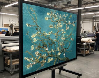 May include: Large framed print of Van Gogh's "Almond Blossoms" against a teal background. The artwork features white blossoms on tree branches. The black frame is displayed on a wheeled stand in a workshop setting.