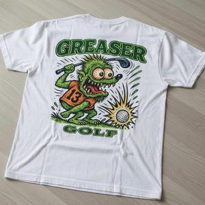 May include: White t-shirt with a cartoon graphic of a green monster playing golf. The monster wears an orange jersey with the number 13. The words "GREASER GOLF" are printed in green.