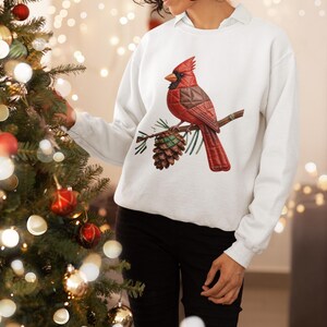 May include: White sweatshirt with a red cardinal bird illustration on a branch with a pinecone. The bird is detailed with a textured design, and the overall aesthetic is festive and seasonal.