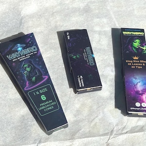 May include: Three boxes of Wayngro rolling papers. The boxes are black with colorful graphics of an alien smoking. One box is labeled "1 1/4 Size" with 6 premium unbleached cones. Another says "40 Leaves + Crutches". The third says "King Size Slim 32 Leaves & 32 Tips".