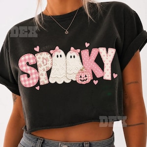 May include: A black cropped t-shirt featuring the word SPOOKY in large, pink and white patterned letters. The letters incorporate two white ghosts with pink bows, a small pink pumpkin, and several small pink hearts scattered around the text.
