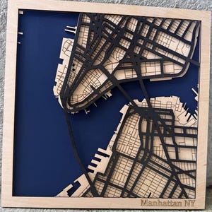 May include: A wooden map of Manhattan, New York, with a dark blue background. The city's street grid is intricately cut out in black, with the words "Manhattan NY" etched at the bottom. The map is framed in light wood.