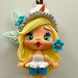 May include: A decorative pen featuring a doll figure with blonde hair, blue eyes, and a white top with blue shorts. The pen has a turquoise gem-studded sphere and a black barrel with doll figures.
