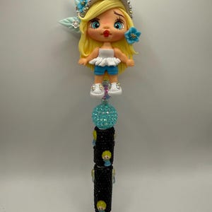 May include: A decorative pen featuring a cartoon doll design. The doll has blonde hair, blue eyes, and is wearing a white top and blue shorts. The pen's barrel is black with small doll accents and a turquoise crystal ball.