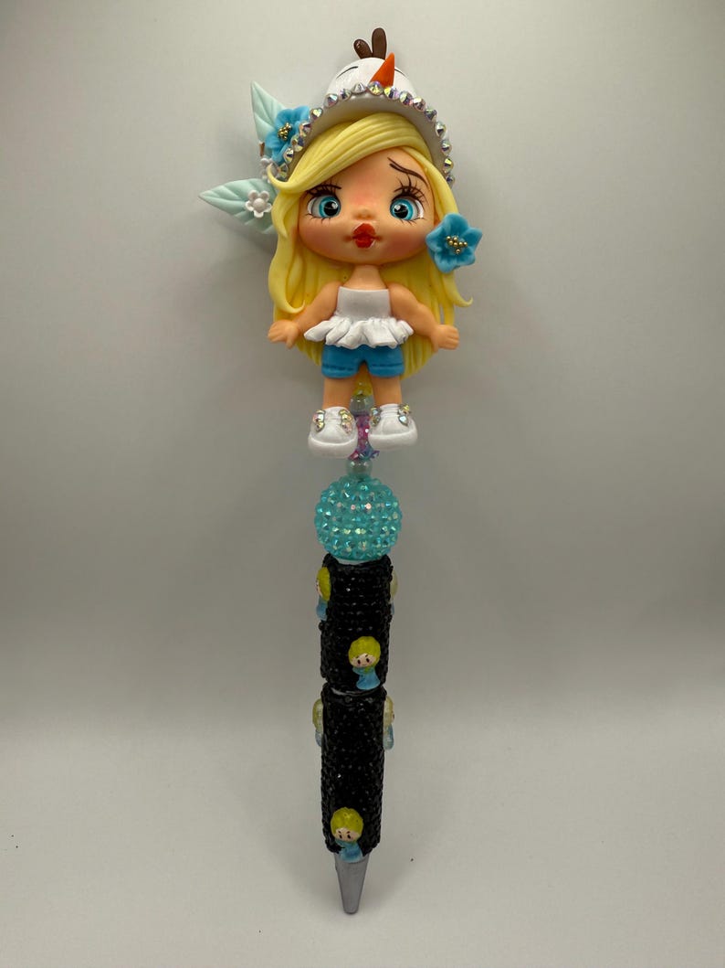 May include: A decorative pen featuring a cartoon doll design. The doll has blonde hair, blue eyes, and is wearing a white top and blue shorts. The pen's barrel is black with small doll accents and a turquoise crystal ball.