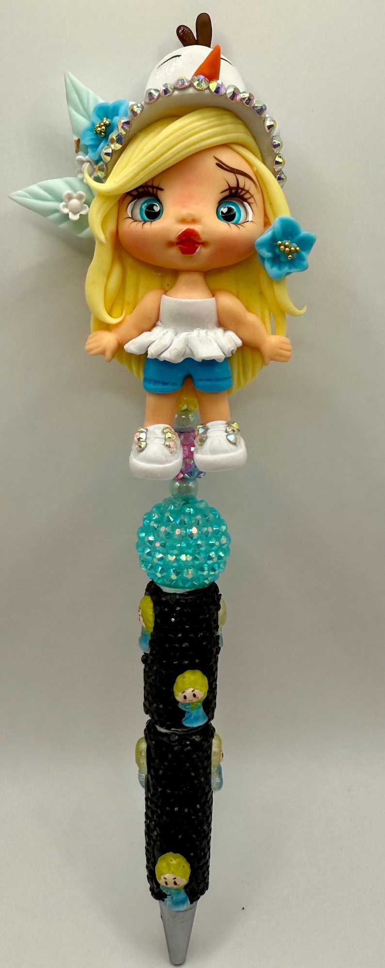 May include: A decorative pen featuring a doll figure with blonde hair, blue eyes, and a white top with blue shorts. The pen has a turquoise gem-studded sphere and a black barrel with doll figures.