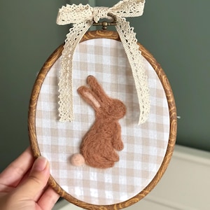 May include: A brown felt bunny is centered on a cream and tan gingham fabric, set within a wooden embroidery hoop. A cream lace bow and ribbon adorn the top. This decorative piece is perfect for Easter or nursery decor.
