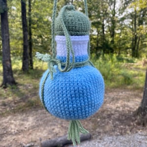 May include: A handmade, blue crochet water bottle holder with a green top and a green tassel. The holder is suspended by a green cord, perfect for outdoor activities. The bottle holder is a unique and stylish accessory.