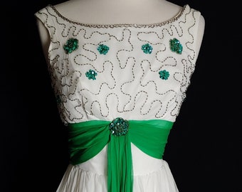 May include: A white sleeveless dress with a green sash and emerald green embellishments. The dress has a fitted bodice and a full skirt.