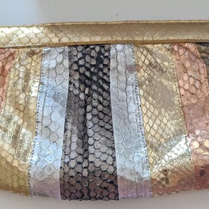 May include: A rectangular clutch with a gold top and a multi-colored snakeskin pattern. The pattern includes vertical stripes of gold, silver, black, and copper tones. The clutch is a stylish accessory.