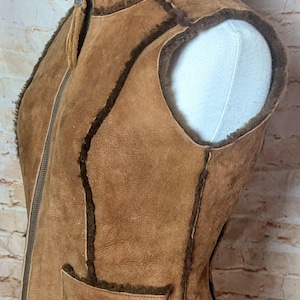 May include: A brown suede vest with a zip-up front and a small pocket. The vest has a dark brown trim and a soft, furry interior. The vest is displayed on a white mannequin.