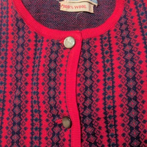 May include: A vintage red wool cardigan with a navy blue geometric pattern. The cardigan features a round neckline, two gold-colored buttons, and a label that reads "100% Wool". The garment is a classic piece of knitwear.