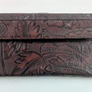 May include: A dark brown leather wallet with a detailed floral and leaf pattern. The wallet has a flap closure and a rectangular shape. The leather appears to be embossed with the design.