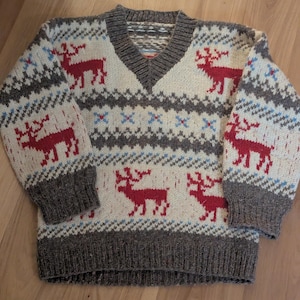 May include: A cream-colored, V-neck sweater with a pattern of red reindeer and geometric shapes. The sweater has gray cuffs, a gray hem, and a gray neckline. Blue accents are also present in the design.