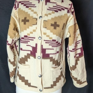 May include: Cream-colored cardigan sweater with a geometric pattern in shades of brown and burgundy. The sweater has a collared neckline, long sleeves, two front pockets, and silver button closures. The pattern includes cross and step-like designs.