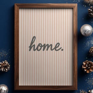Op de afbeelding: Een houten lijst met het woord "home" in een sierlijk lettertype. De achtergrond heeft verticale beige en witte strepen. De lijst is donkerbruin. Denappels en zilveren ballen omringen de lijst op een donkerblauwe ondergrond, wat een feestelijke sfeer creëert.