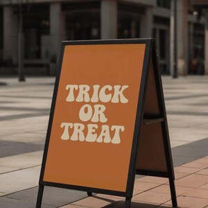 Puede incluir: Un letrero naranja con forma de caballete con las palabras "TRICK OR TREAT" en una fuente retro. El letrero tiene un marco de metal negro y se muestra al aire libre sobre una superficie de ladrillo. El letrero es probablemente para Halloween.