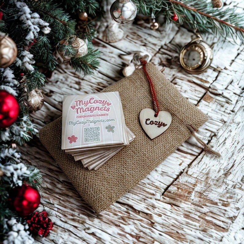 K&ouml;nnte beinhalten: Ein Stapel quadratischer Magnete mit dem Text "My Cozy Magnets" und einer Website-Adresse, auf einem Jutebeutel. Ein Holzherz mit dem Wort "Cozy" h&auml;ngt an der Tasche. Weihnachtsdekorationen, darunter Ornamente und eine Uhr, sind im Hintergrund.