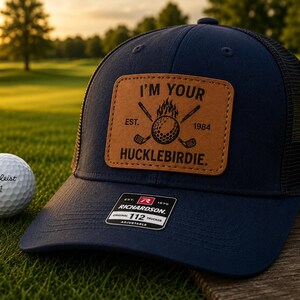 Peut inclure: Une casquette trucker bleu marine avec un filet noir à l'arrière et un écusson en cuir beige sur le devant. L'écusson présente une balle de golf enflammée, des clubs croisés et le texte I'M YOUR HUCKLEBIRDIE. EST. 1984. Une balle de golf Titleist 1 est posée à côté.