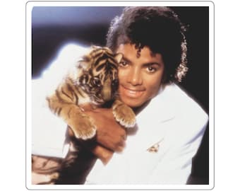 May include: A sticker featuring a portrait of Michael Jackson wearing a white blazer, smiling while holding a small tiger cub with orange and black stripes. The image has a dark background and a white border, capturing a clear, detailed view.