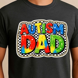May include: Dark gray t-shirt with the text "AUTISM DAD" in colorful, bubble letters. The text is outlined in black and set against a checkered background. The shirt is a crew neck style.
