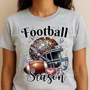 Könnte beinhalten: Graues T-Shirt mit einem Football-Motiv. Das Design umfasst einen Helm mit Leopardenmuster, einen braunen Football und die Worte "Football Season". Glitzernde Edelsteine und ein Herz vervollständigen das Design.