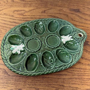 May include: Green ceramic egg serving platter with oval shape and basket weave design. Features 10 egg-shaped indentations and two round indentations. Decorated with white floral accents and a handle with a hole.