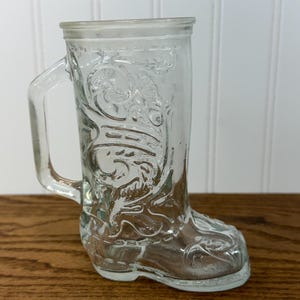May include: Clear glass boot-shaped mug with a handle. The mug features embossed decorative patterns and is set on a wooden surface. The boot design includes details like a pointed toe and a decorative top.