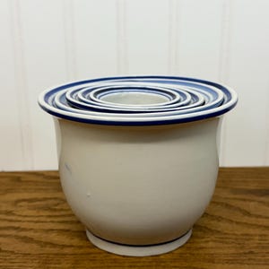 May include: A set of white ceramic measuring bowls with a blue rim and accents. The bowls are nested inside each other, with the largest bowl at the bottom and the smallest at the top. The bowls are sitting on a wooden surface.