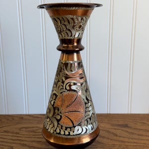 May include: A copper-toned vase with intricate floral and leaf patterns in silver and gold. The vase has a flared top and a conical body, with a round base. The design is etched into the metal, creating a detailed, decorative piece.