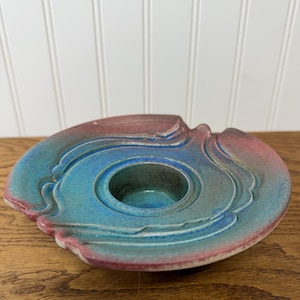 May include: A ceramic candle holder with a teal and red color scheme. The holder has a circular design with a central well for a candle and layered, wave-like edges. The piece is on a wooden surface.