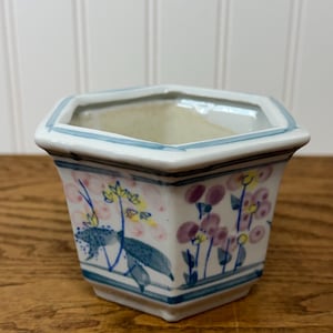 May include: A hexagonal ceramic planter with a white base, featuring hand-painted floral motifs in shades of pink, yellow, and blue. The rim and base are accented with thin blue horizontal lines, and the interior is a plain, off-white glazed finish.