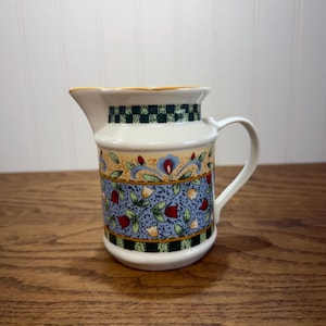 May include: A ceramic creamer pitcher featuring a cream-colored base with a decorative band of blue, red, and yellow floral patterns. The rim and base are accented with a black and green checkered border and a curved handle on the side.