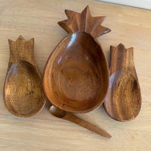 May include: A wooden pineapple-shaped bowl with a small spoon and two spoon-shaped serving utensils. The set is made of a light brown wood and is arranged on a light-colored wooden surface. The bowl has a star-shaped top.