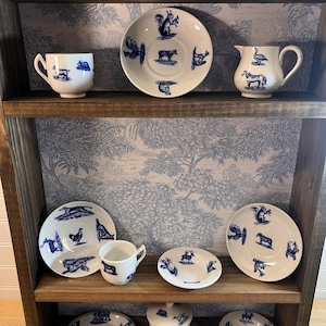 May include: A wooden shelf displays a collection of white ceramic dishware with blue animal illustrations. The set includes cups, plates, a creamer, and a covered dish. The shelf is against a blue and white patterned wallpaper.