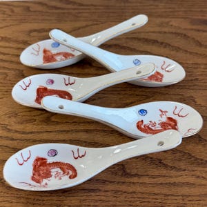 May include: Set of four white ceramic soup spoons. Each spoon features a red and gold dragon design with gold trim. The spoons are arranged on a wooden surface.