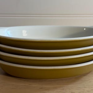 May include: A stack of four oval-shaped serving dishes. The dishes have a mustard yellow exterior and a white interior. Each dish has a handle on either side. The dishes are stacked on top of each other.