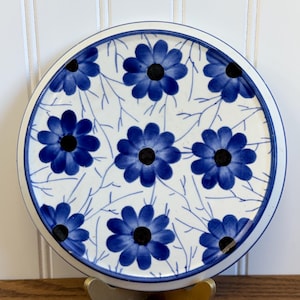 May include: A round, white ceramic coaster with a blue floral design. The coaster features blue flowers with black centers and delicate blue leaf patterns. The edge is trimmed with a blue border. The coaster is displayed on a gold-colored stand.