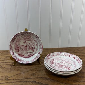 May include: A set of small round ceramic saucers featuring a maroon transferware pattern on a white background. The design depicts figures in a landscape with buildings and a boat. One saucer is displayed on a gold metal stand.