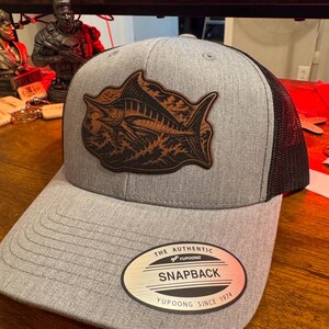 May include: A heather grey snapback cap with a black mesh back and a brown leather patch featuring a detailed marlin fish graphic. The brim displays a circular sticker with the text THE AUTHENTIC YUPOONG SNAPBACK YUPOONG SINCE 1974.