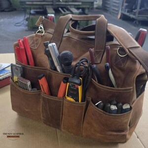 Puede incluir: Una bolsa de herramientas de cuero marrón con múltiples bolsillos llenos de varias herramientas. La bolsa tiene un asa y una correa para el hombro. Las herramientas incluyen alicates, destornilladores y un multímetro amarillo. La bolsa está etiquetada como "TRENDY LEATHER GIFTS".