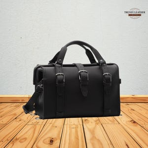 May include: Black leather satchel bag with a top handle and adjustable shoulder strap. The bag features three buckle straps on the front and a flap closure. The bag is sitting on a wooden surface.
